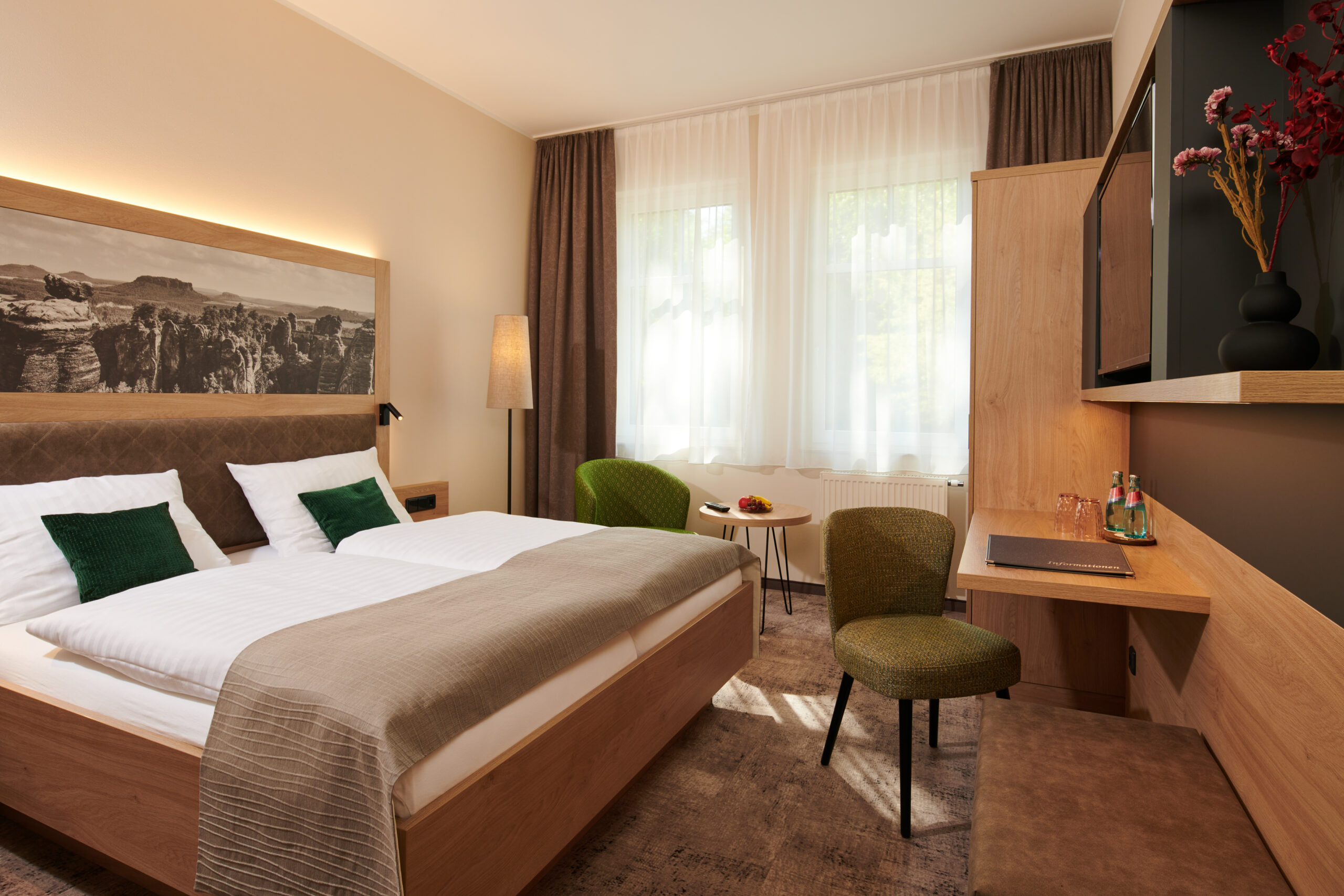 Elegantes Hotelzimmer im Parkhotel Neustadt, ausgestattet mit einem komfortablen Bett mit weißer Bettwäsche und grünen Akzentkissen, einer Sitzecke mit grünen Stühlen, einem Schreibtisch und einem flachen Fernseher auf einem Regal. Das Zimmer ist mit warmen Brauntönen und sanfter Beleuchtung gestaltet, was eine einladende Atmosphäre schafft.